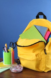 Backpack with different school stationery on wooden table against blue background