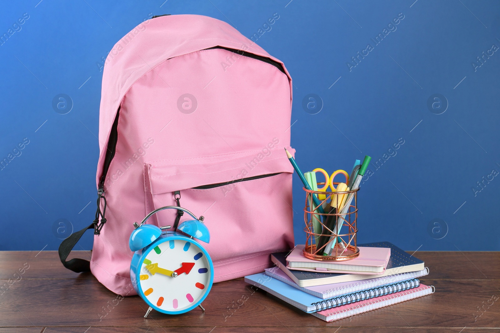 Photo of Backpack with different school stationery and alarm clock on wooden table against blue background