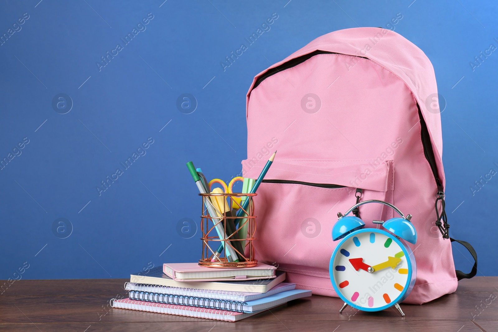 Photo of Backpack with different school stationery and alarm clock on wooden table against blue background, space for text