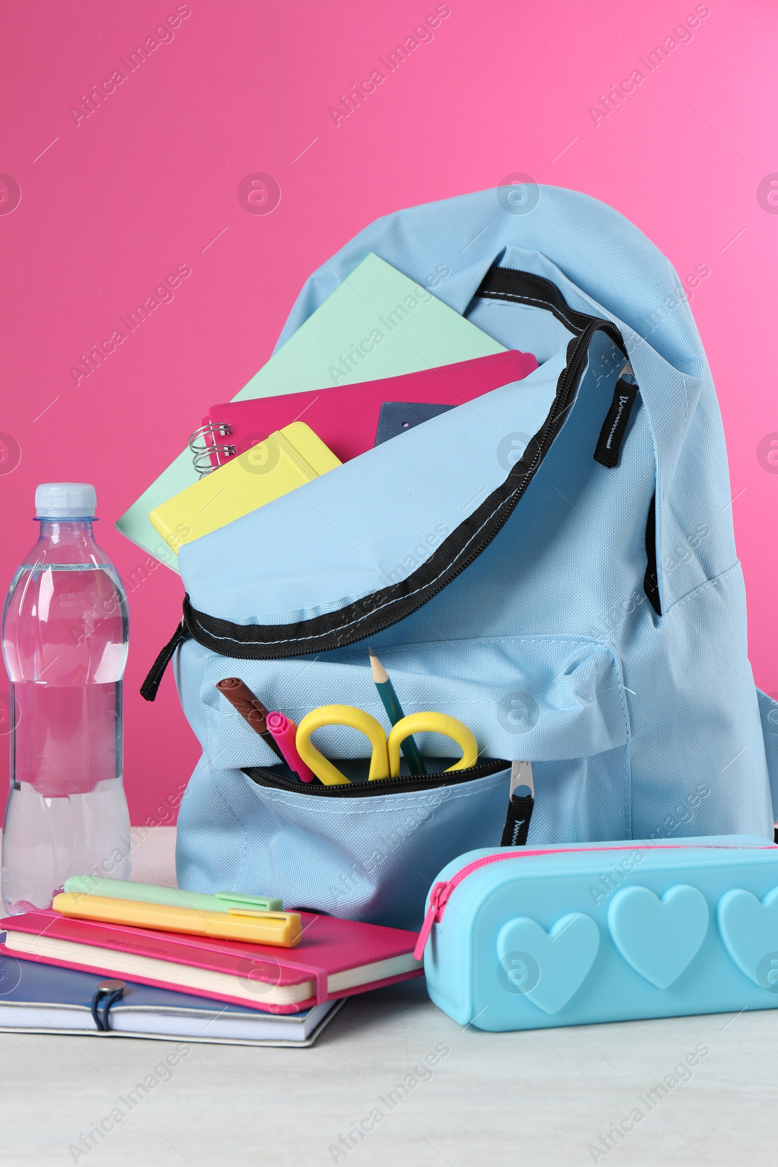 Photo of Backpack with different school stationery and bottle of water on light table against pink background