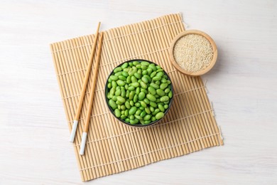 Fresh edamame soybeans in bowl, chopsticks and sesame seeds on white wooden table, top view