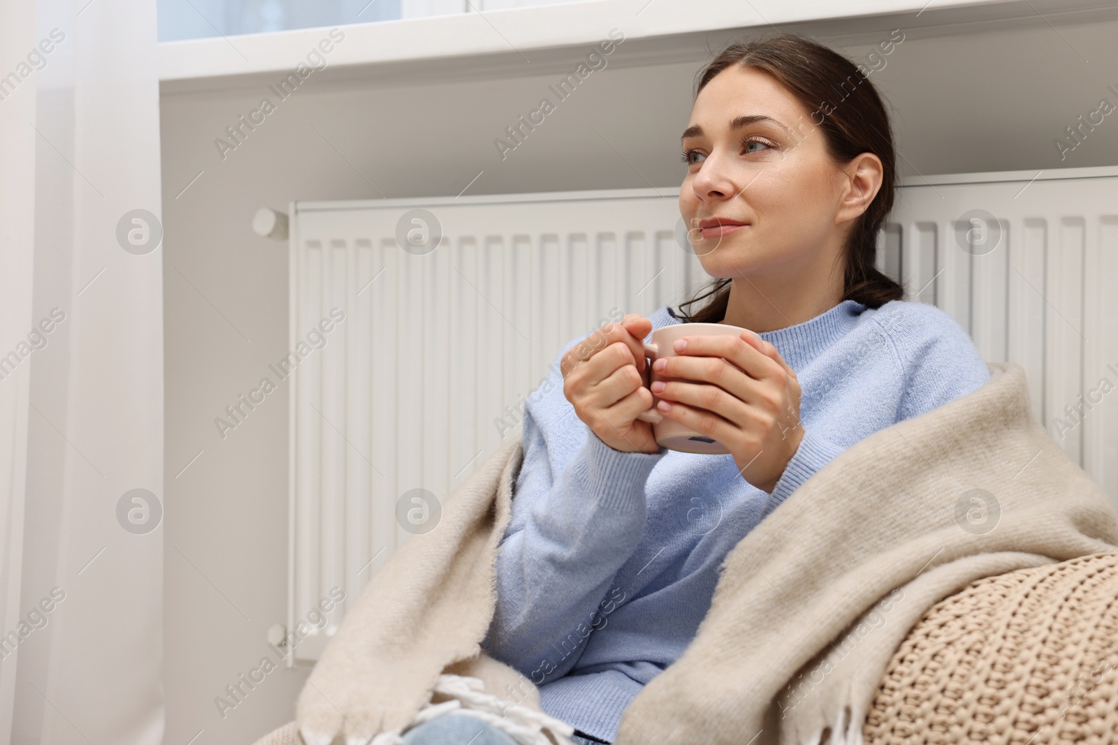 Photo of Woman with cup of hot drink near heating radiator at home