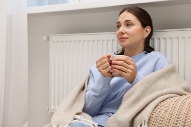 Woman with cup of hot drink near heating radiator at home