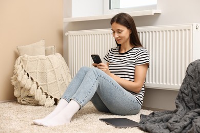 Happy woman using smartphone near heating radiator at home