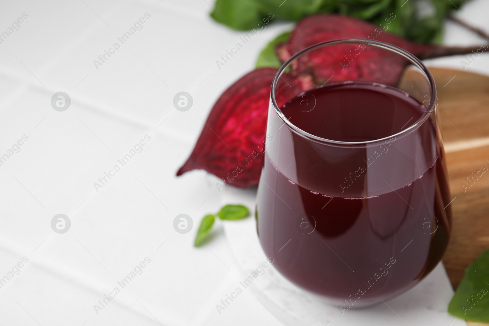 Photo of Fresh beet juice in glass and ripe vegetables on white tiled table, closeup. Space for text