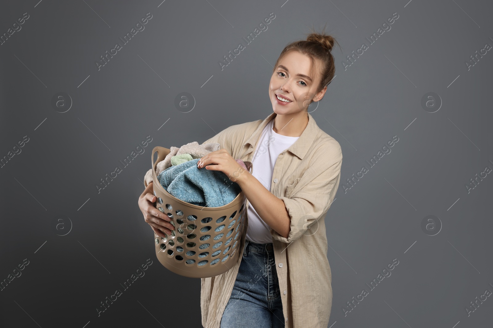 Photo of Happy young housewife with basket full of laundry on grey background