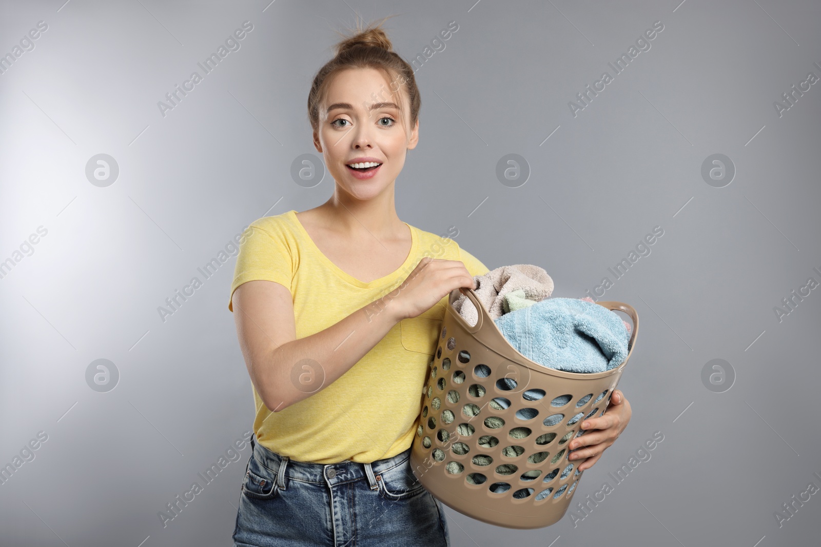 Photo of Happy young housewife with basket full of laundry on grey background