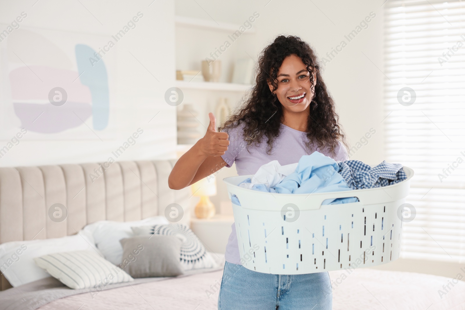 Photo of Happy woman with basket full of laundry showing thumbs up in bedroom, space for text