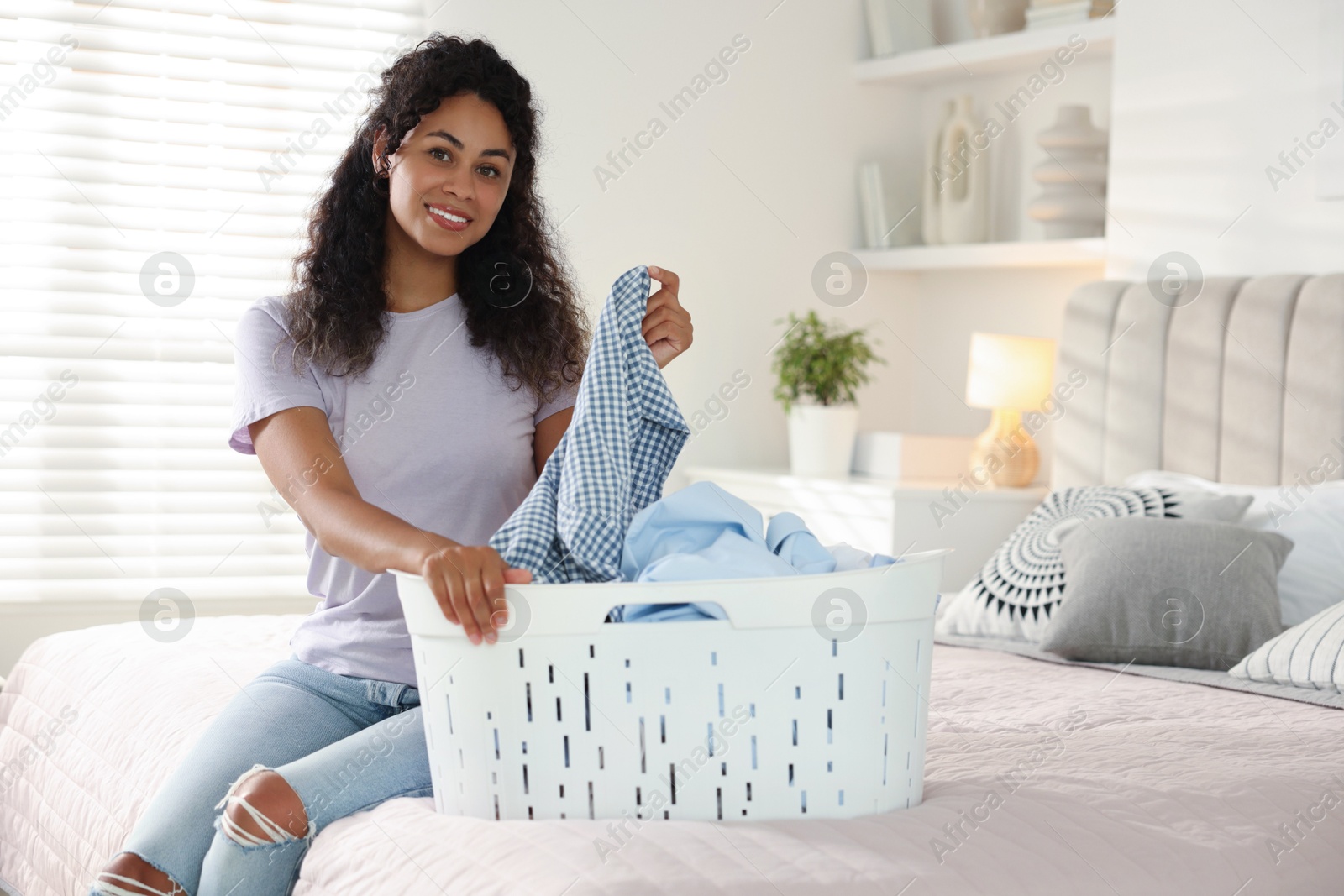 Photo of Happy woman with basket full of laundry on bed at home