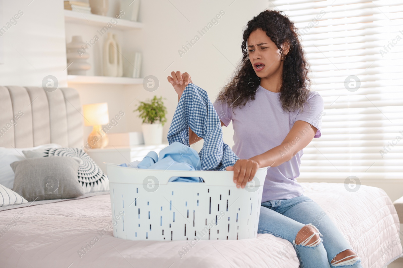 Photo of Displeased woman with basket full of laundry on bed at home