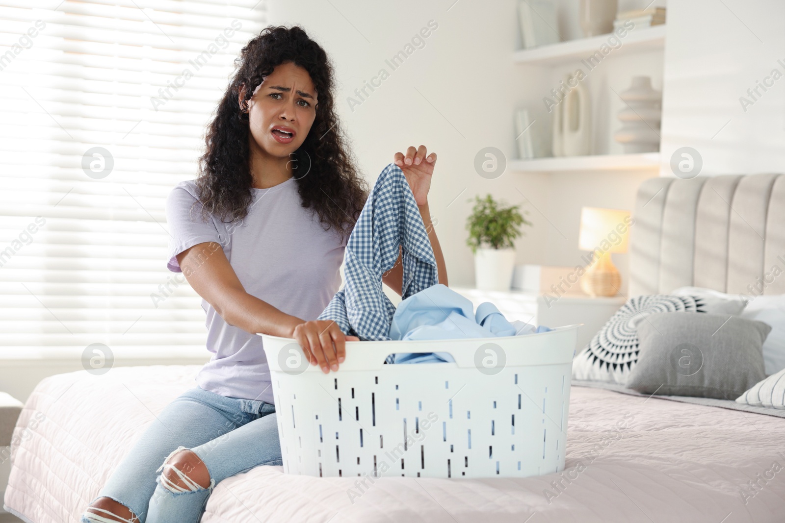 Photo of Displeased woman with basket full of laundry on bed at home