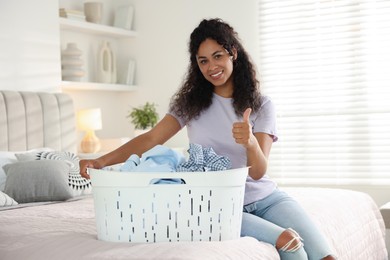 Happy woman with basket full of laundry showing thumbs up on bed at home