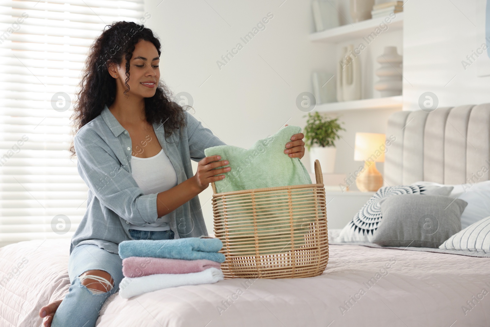 Photo of Happy woman with basket full of laundry on bed at home