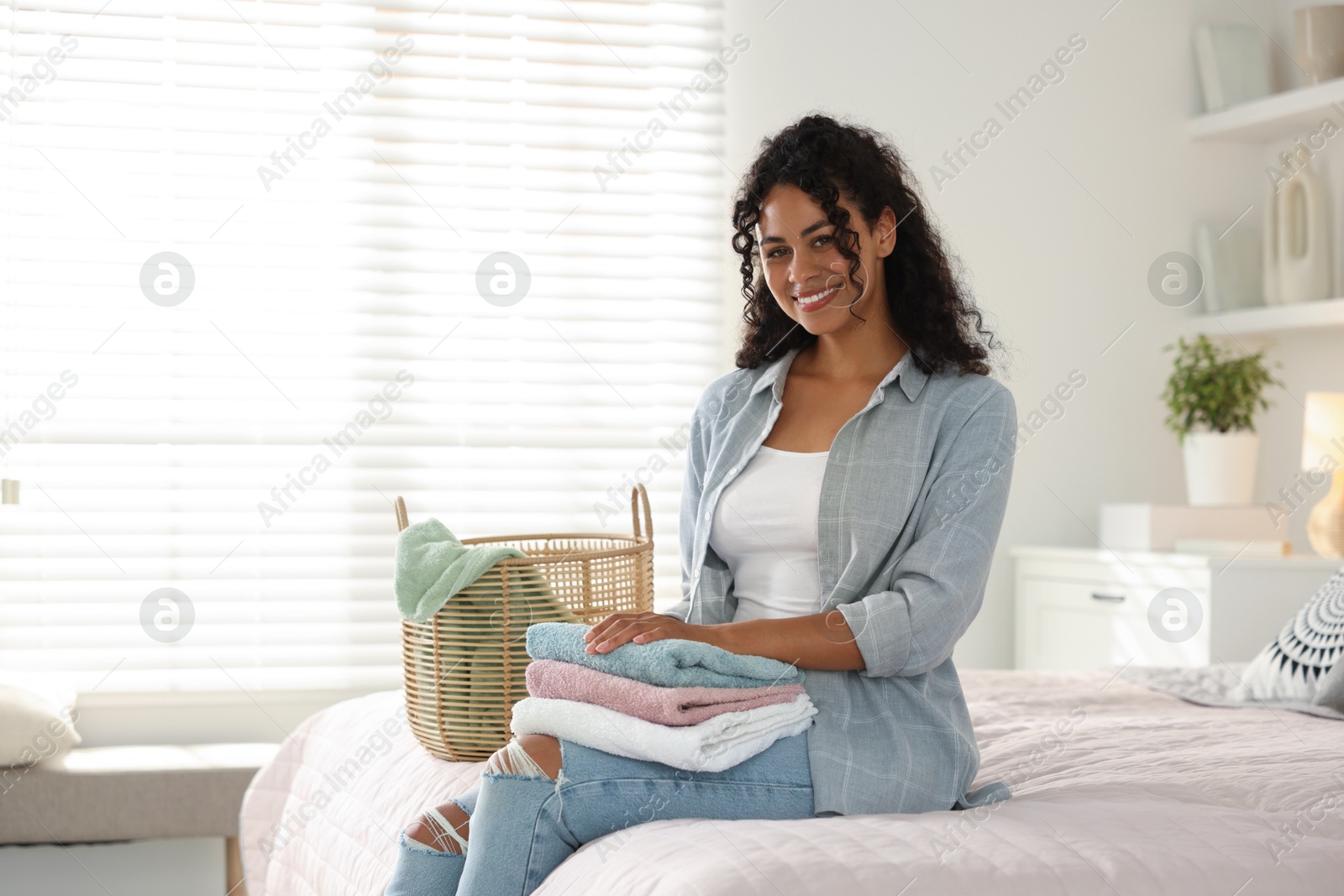 Photo of Happy woman with laundry near basket on bed at home