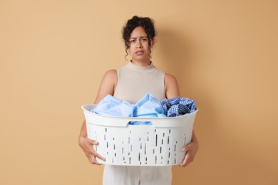 Displeased woman with basket full of laundry on beige background