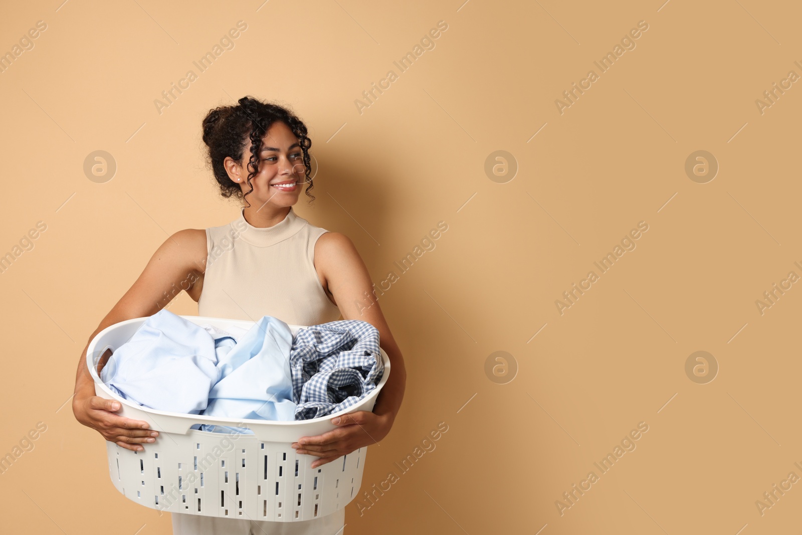 Photo of Happy woman with basket full of laundry on beige background, space for text