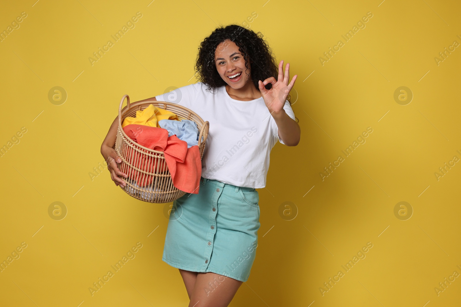 Photo of Happy woman with basket full of laundry showing ok gesture on yellow background