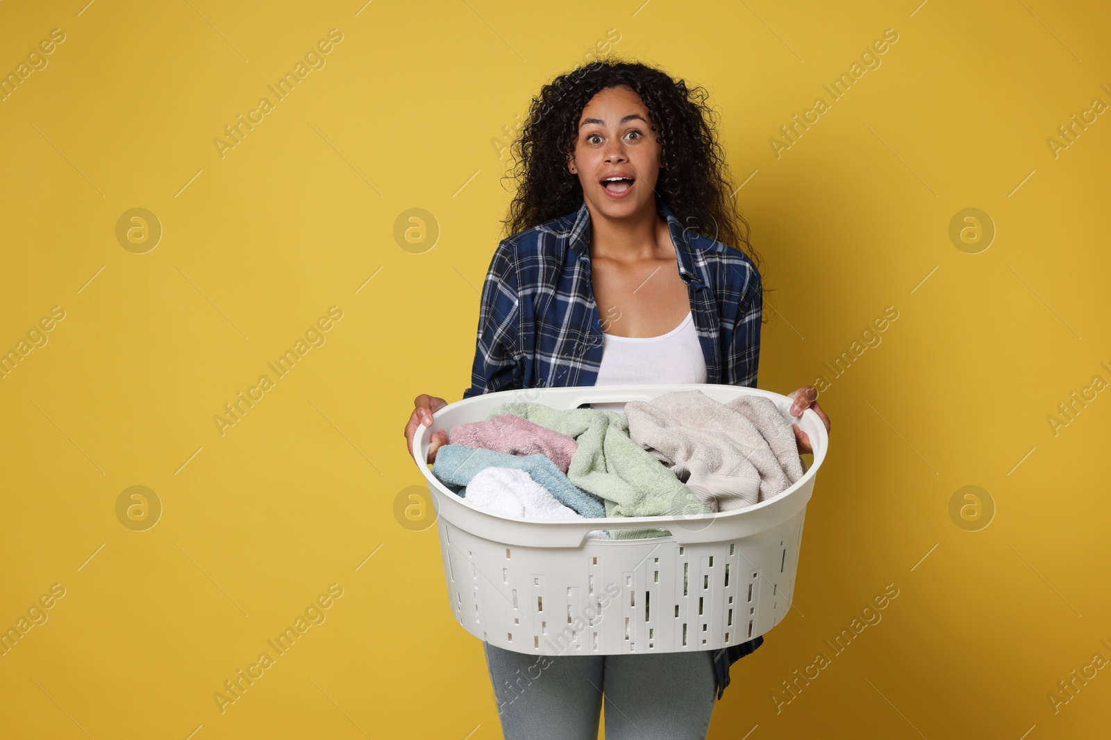 Photo of Shocked woman with basket full of laundry on yellow background, space for text