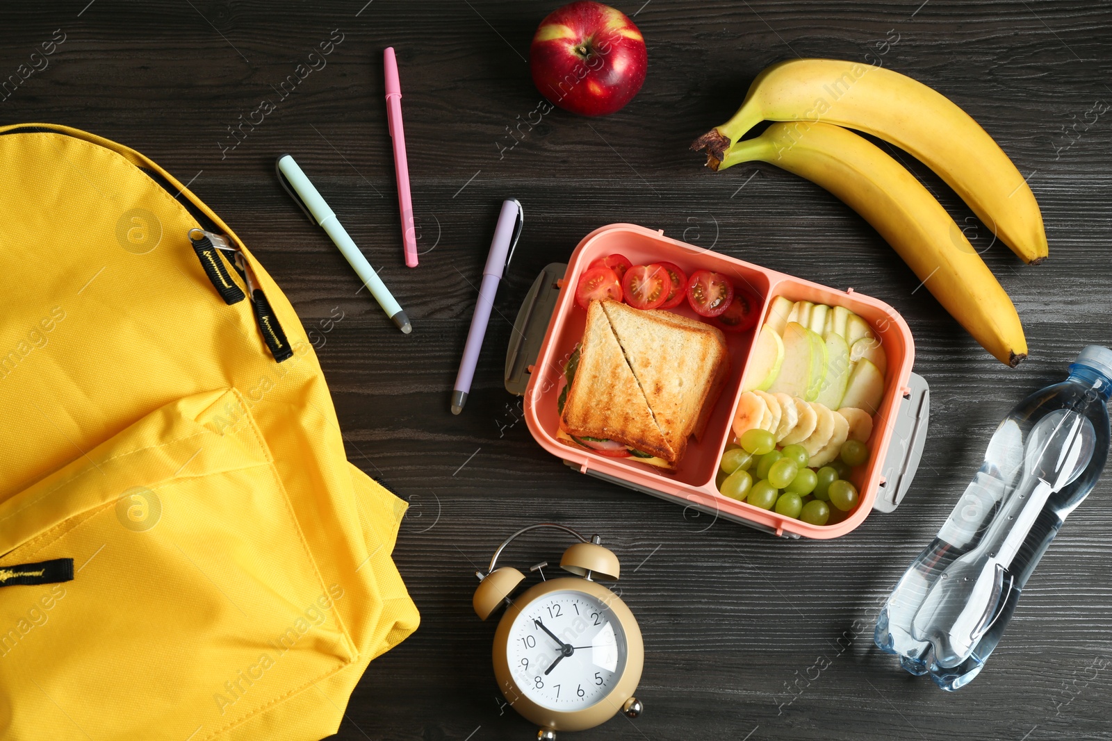 Photo of Backpack, lunch box with snacks, bottle of water and alarm clock on wooden table, flat lay