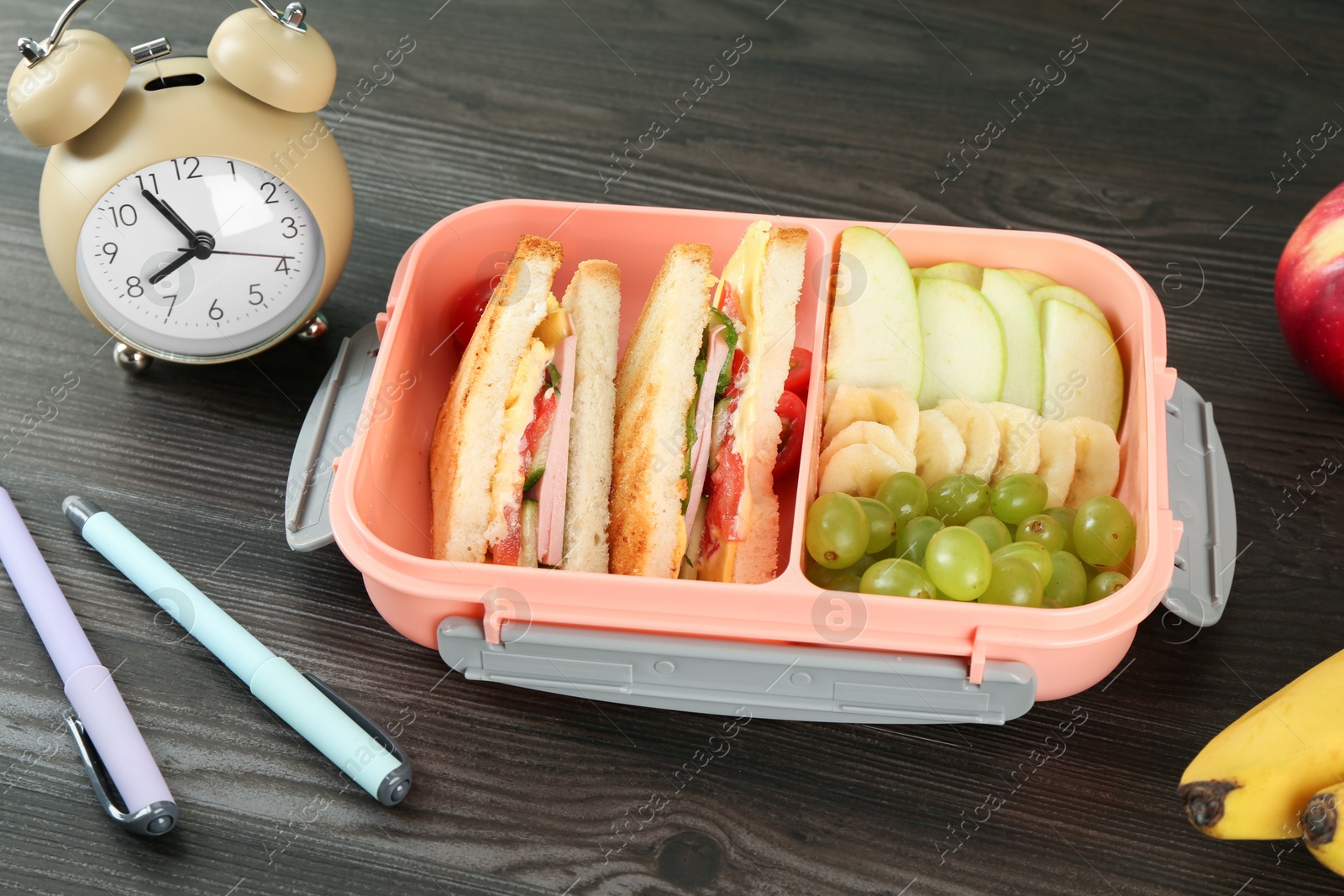 Photo of Lunch box with snacks, alarm clock and pens on wooden table