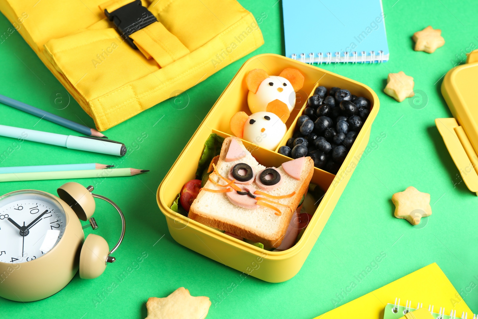 Photo of Bag, lunch box with snacks, stationery and alarm clock on green table, closeup