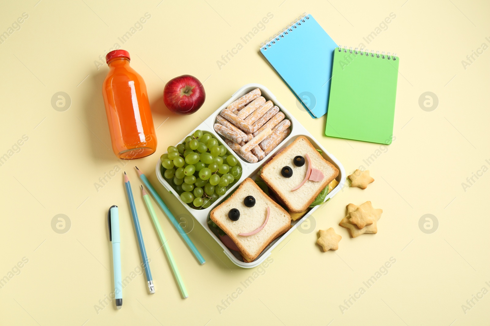 Photo of Sandwiches with smiley faces in lunch box, bottle of juice and stationery on yellow table, flat lay