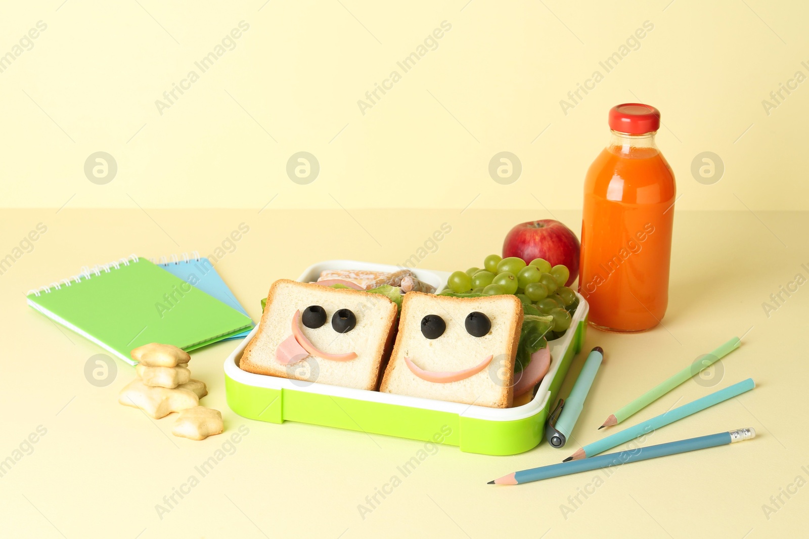 Photo of Sandwiches with smiley faces in lunch box, bottle of juice and stationery on yellow table