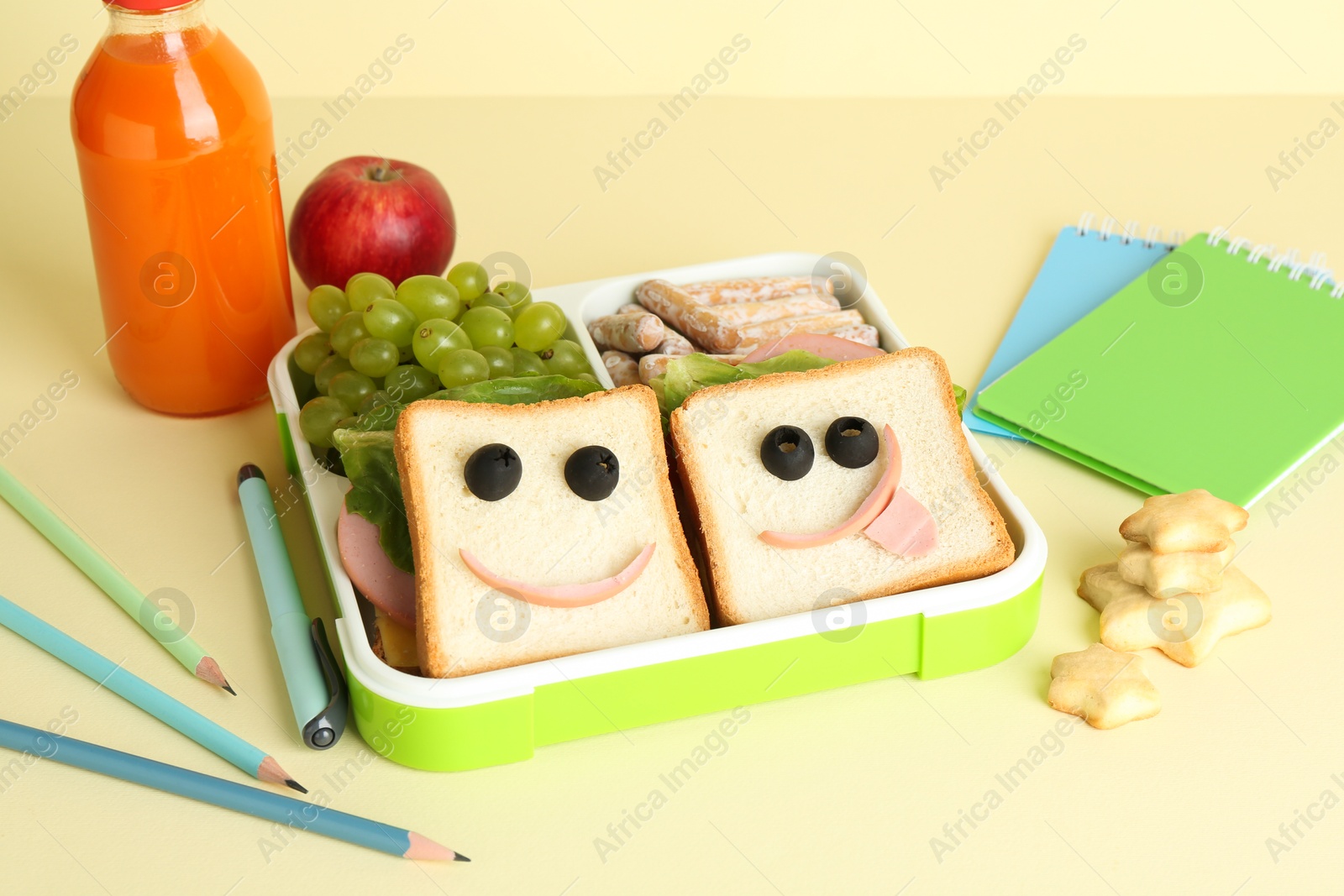 Photo of Sandwiches with smiley faces in lunch box, bottle of juice and stationery on yellow table