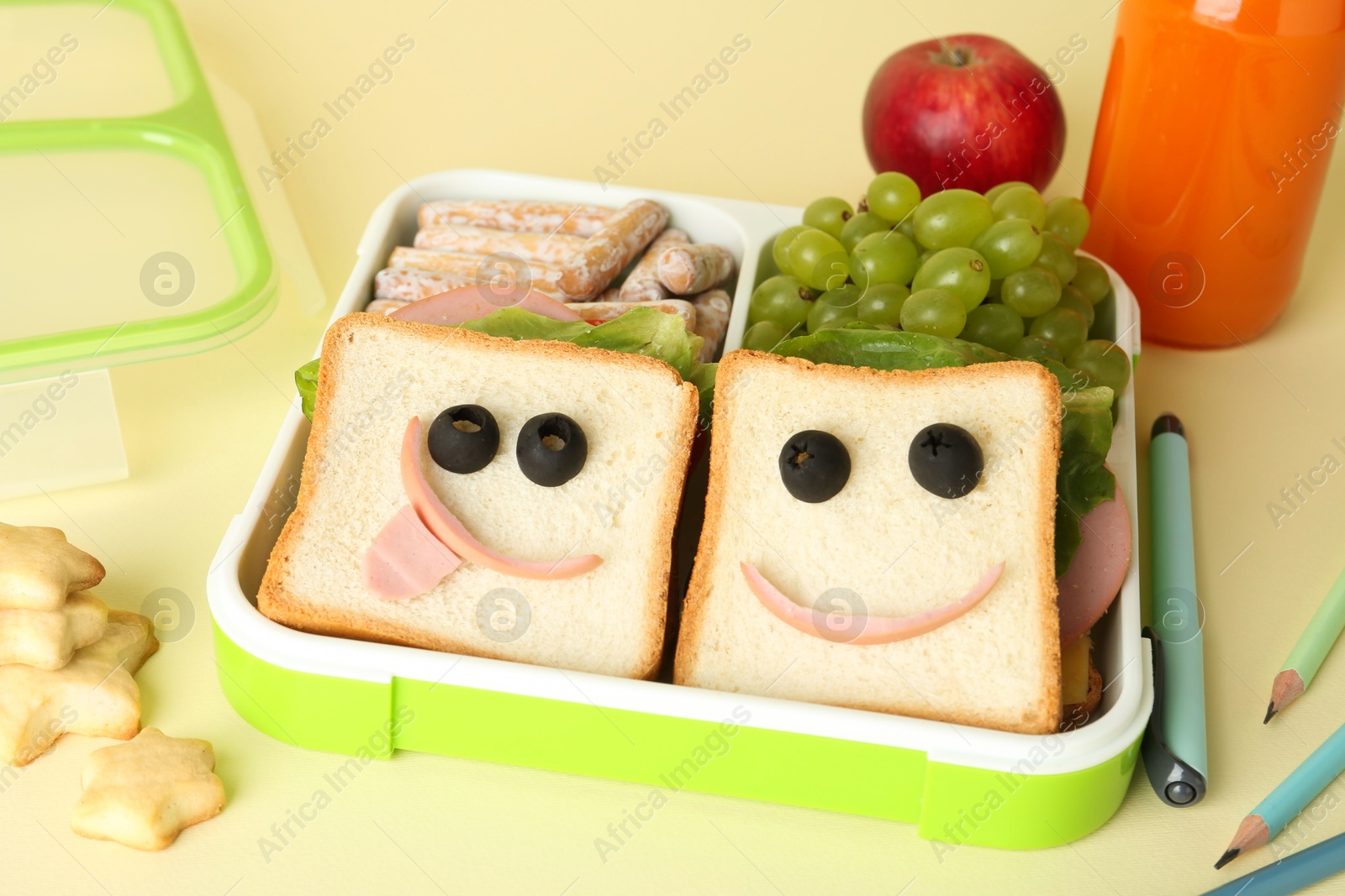 Photo of Sandwiches with smiley faces in lunch box, bottle of juice, pencils and pen on yellow table, closeup