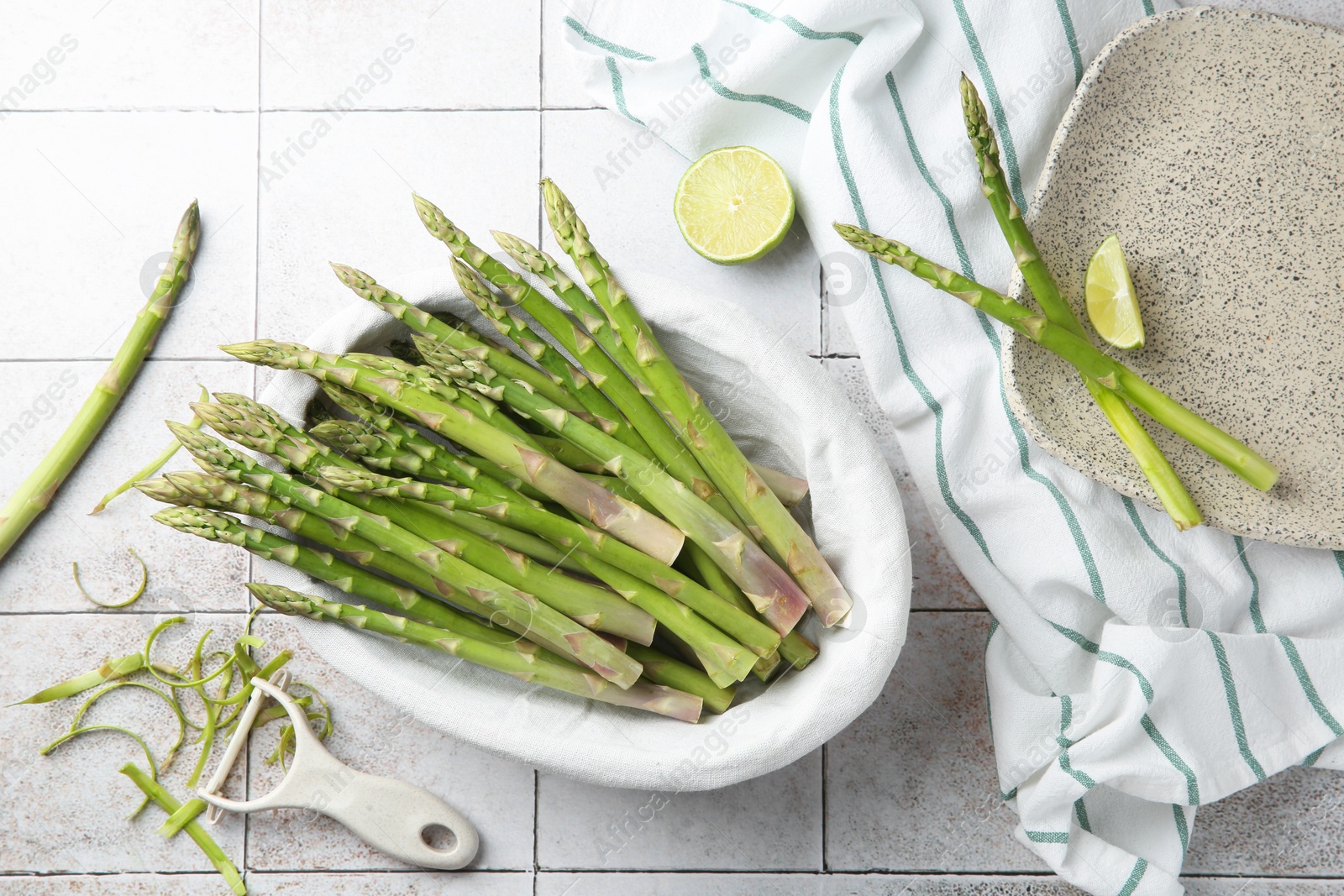 Photo of Fresh green asparagus stems, lime and vegetable peeler on light textured tiled table, flat lay