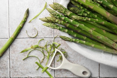 Fresh green asparagus stems and vegetable peeler on light textured tiled table, flat lay