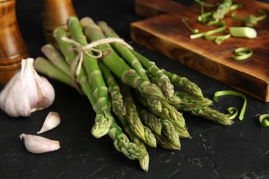 Photo of Fresh green asparagus stems and garlic on black textured table, closeup