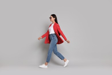 Photo of Stylish woman in red jacket, jeans and sunglasses walking on gray background