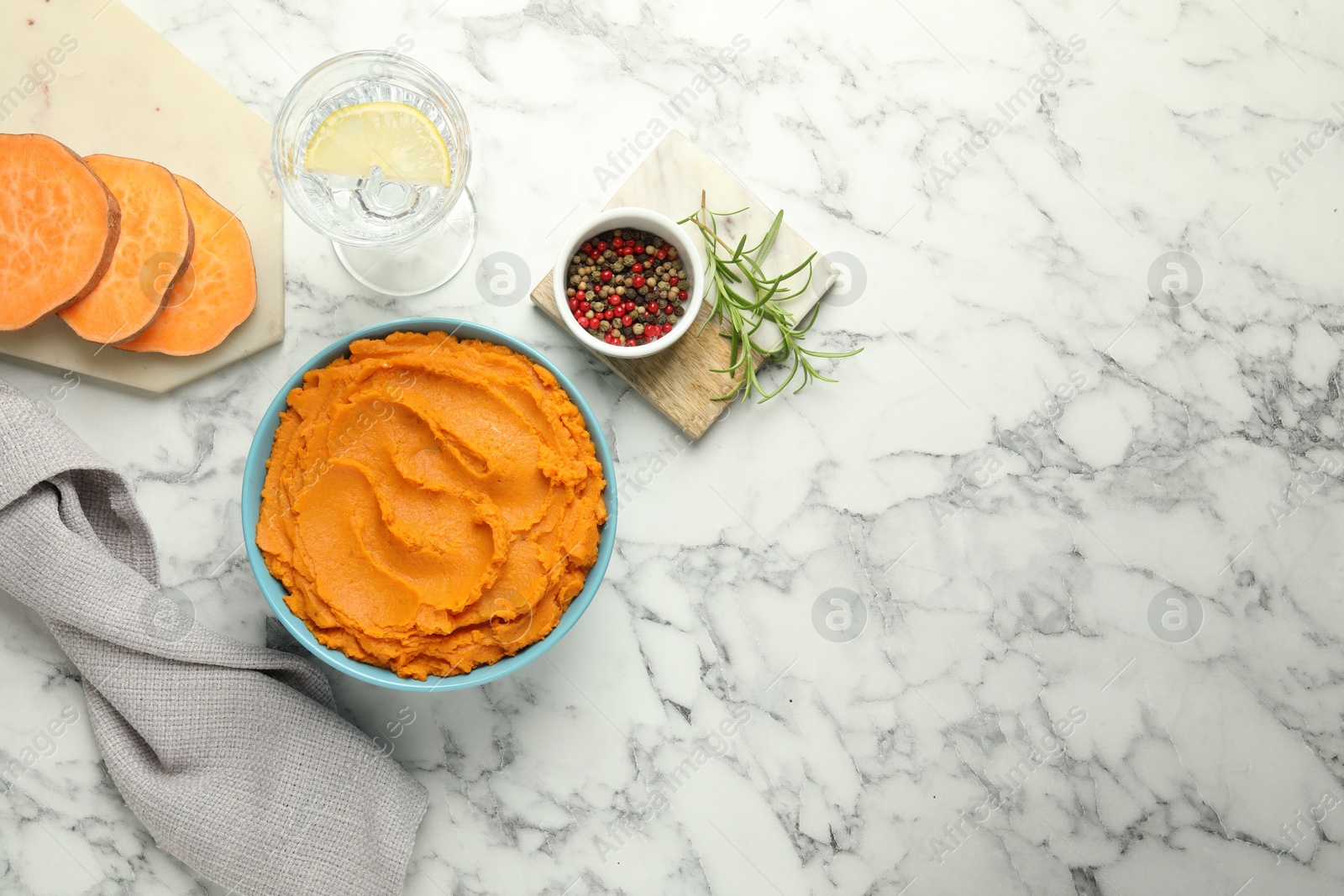 Photo of Tasty mashed sweet potato in bowl, peppercorns and water with lemon on white marble table, flat lay. Space for text