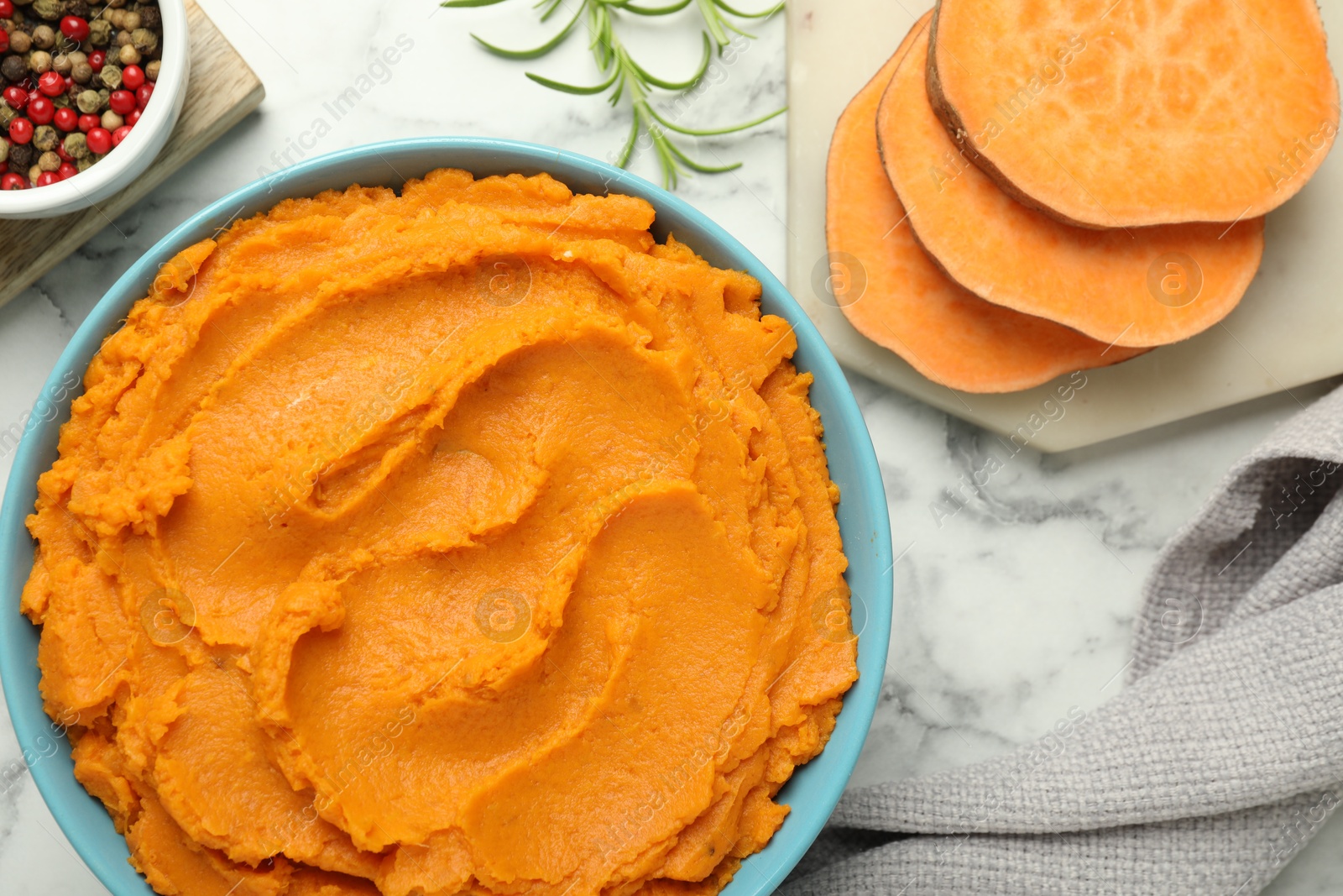 Photo of Tasty mashed sweet potato in bowl, peppercorns and rosemary on white marble table, flat lay