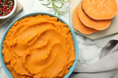 Tasty mashed sweet potato in bowl, peppercorns and rosemary on white marble table, flat lay