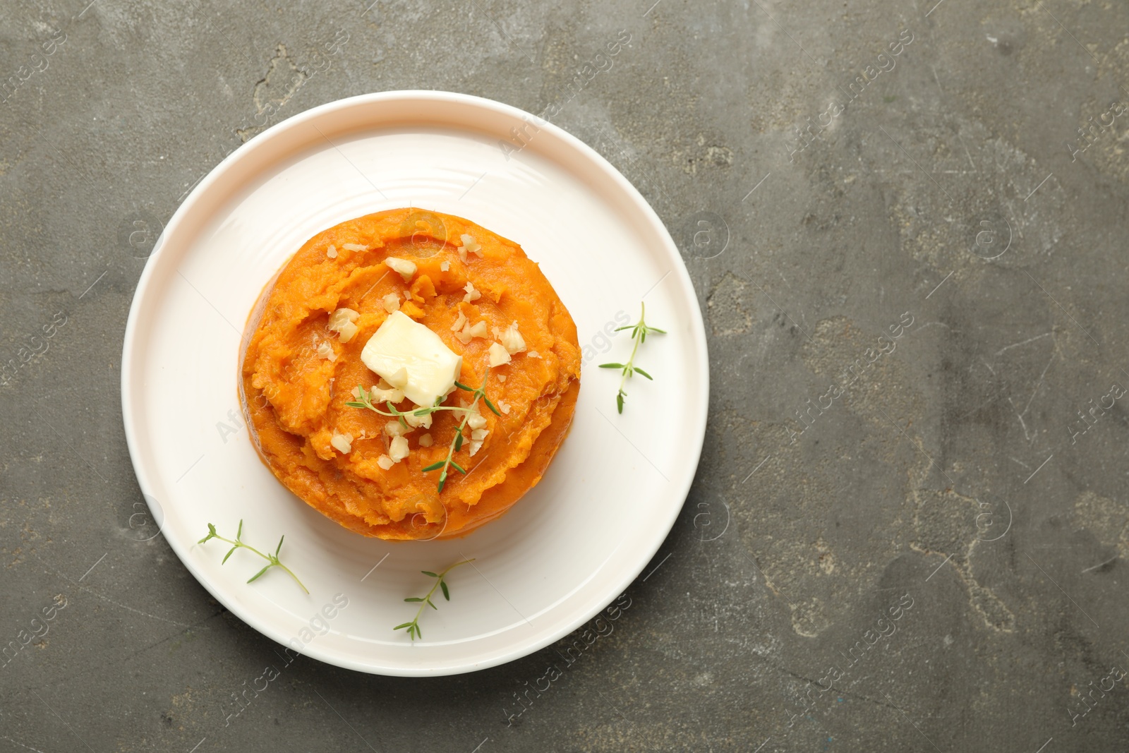 Photo of Plate with tasty mashed sweet potato, butter, thyme and walnuts on grey textured table, top view. Space for text