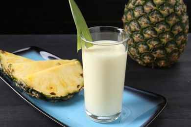 Tasty pineapple smoothie in glass and fruit on black wooden table, closeup