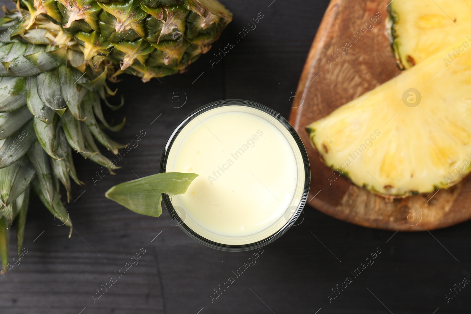 Photo of Tasty pineapple smoothie in glass and slices of fruit on black wooden table, flat lay