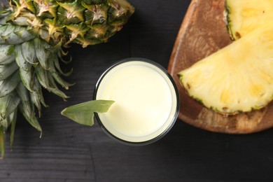 Photo of Tasty pineapple smoothie in glass and slices of fruit on black wooden table, flat lay