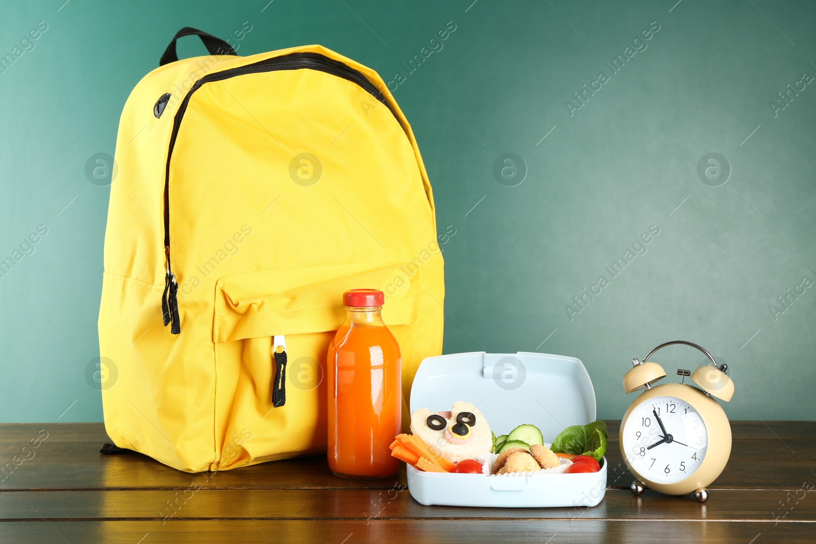 Photo of Backpack, lunch box with snacks, bottle of juice and alarm clock on wooden table against color background