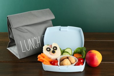 Photo of Gray bag, lunch box with sandwich, fresh vegetables, cookies and peach on wooden table against color background