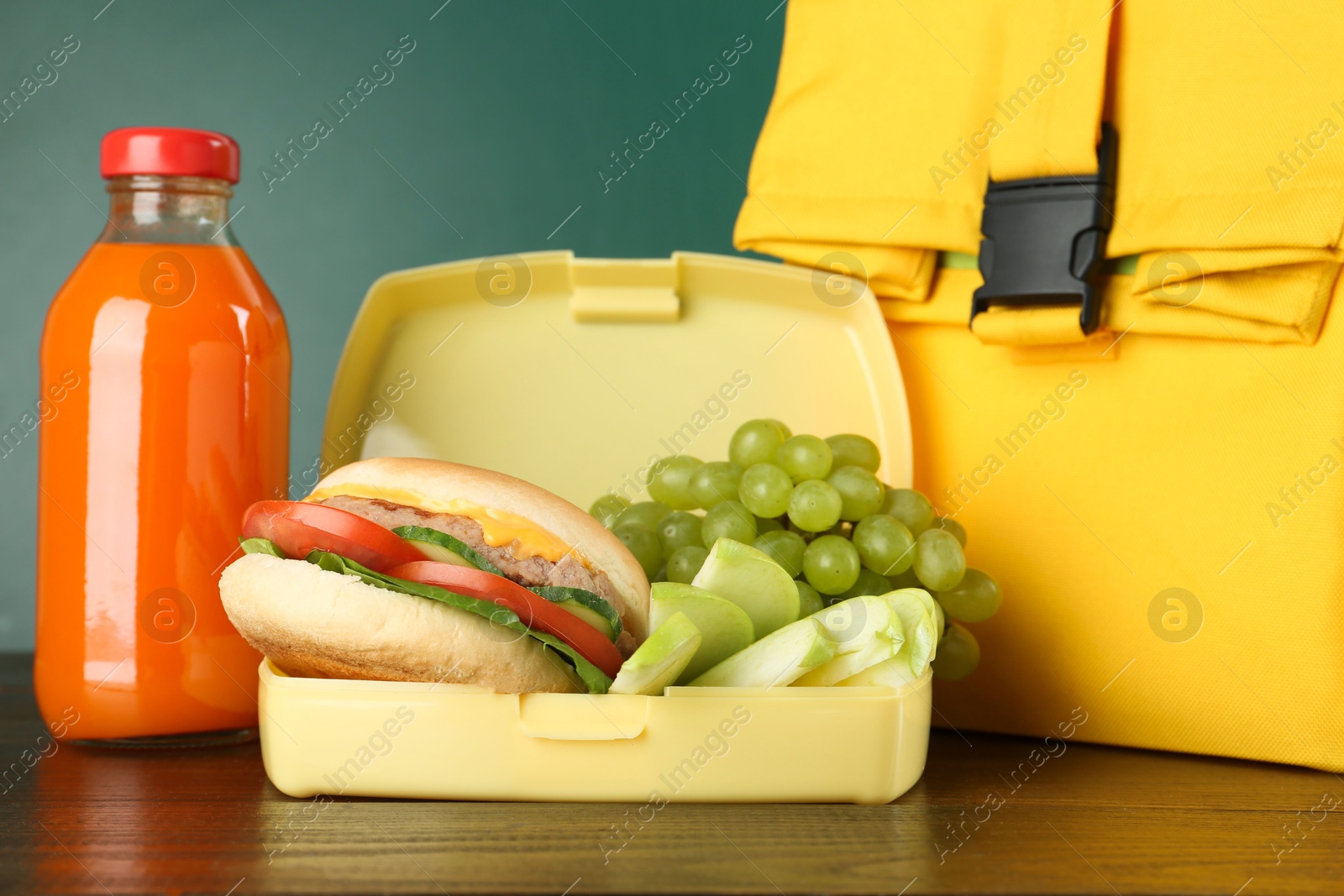 Photo of Bag, lunch box with burger, fruits and bottle of juice on wooden table against color background, closeup