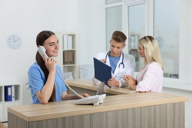 Professional receptionist and doctor working with patient at wooden desk in hospital