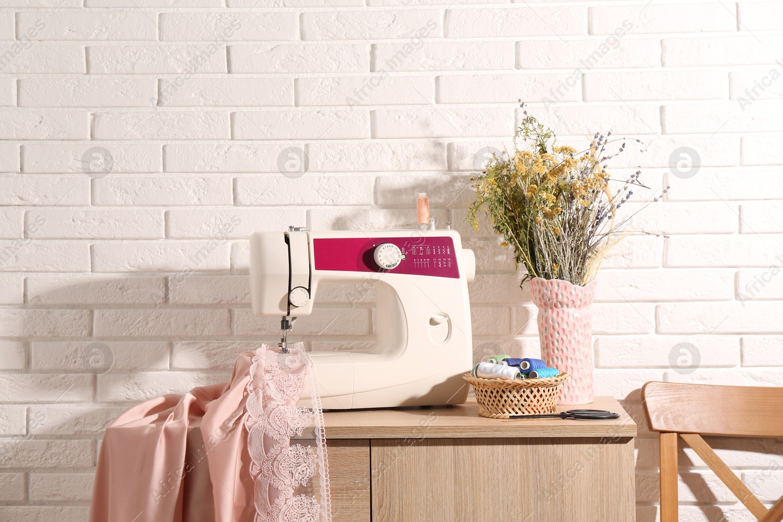 Photo of Sewing machine with fabric, flowers in vase and tools on chest of drawers indoors