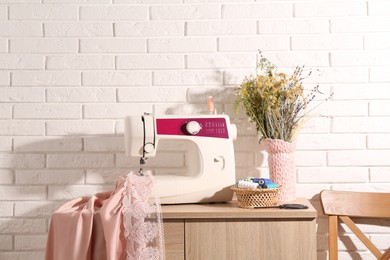 Photo of Sewing machine with fabric, flowers in vase and tools on chest of drawers indoors