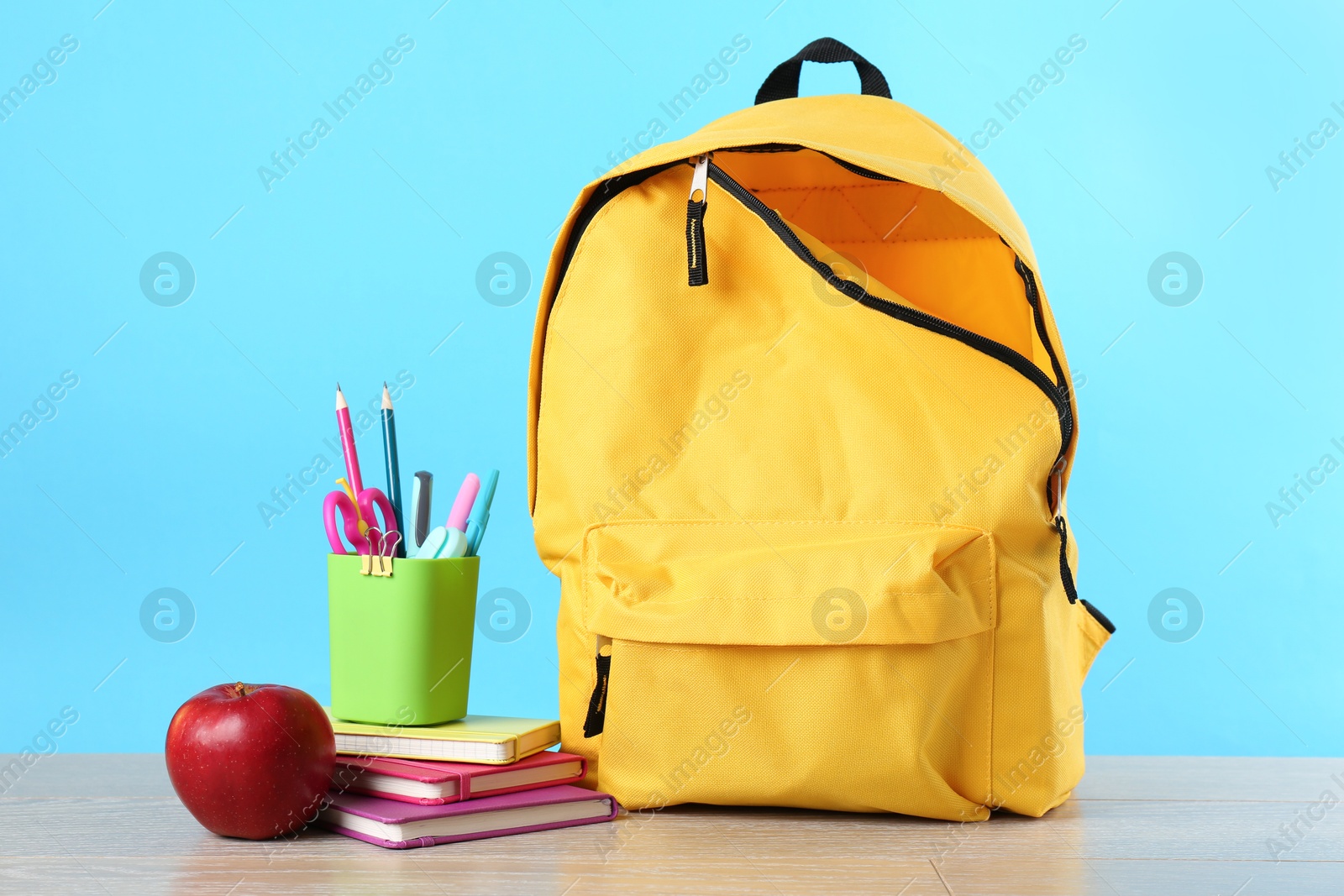 Photo of Backpack with different school stationery and apple on wooden table against light blue background