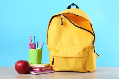 Backpack with different school stationery and apple on wooden table against light blue background