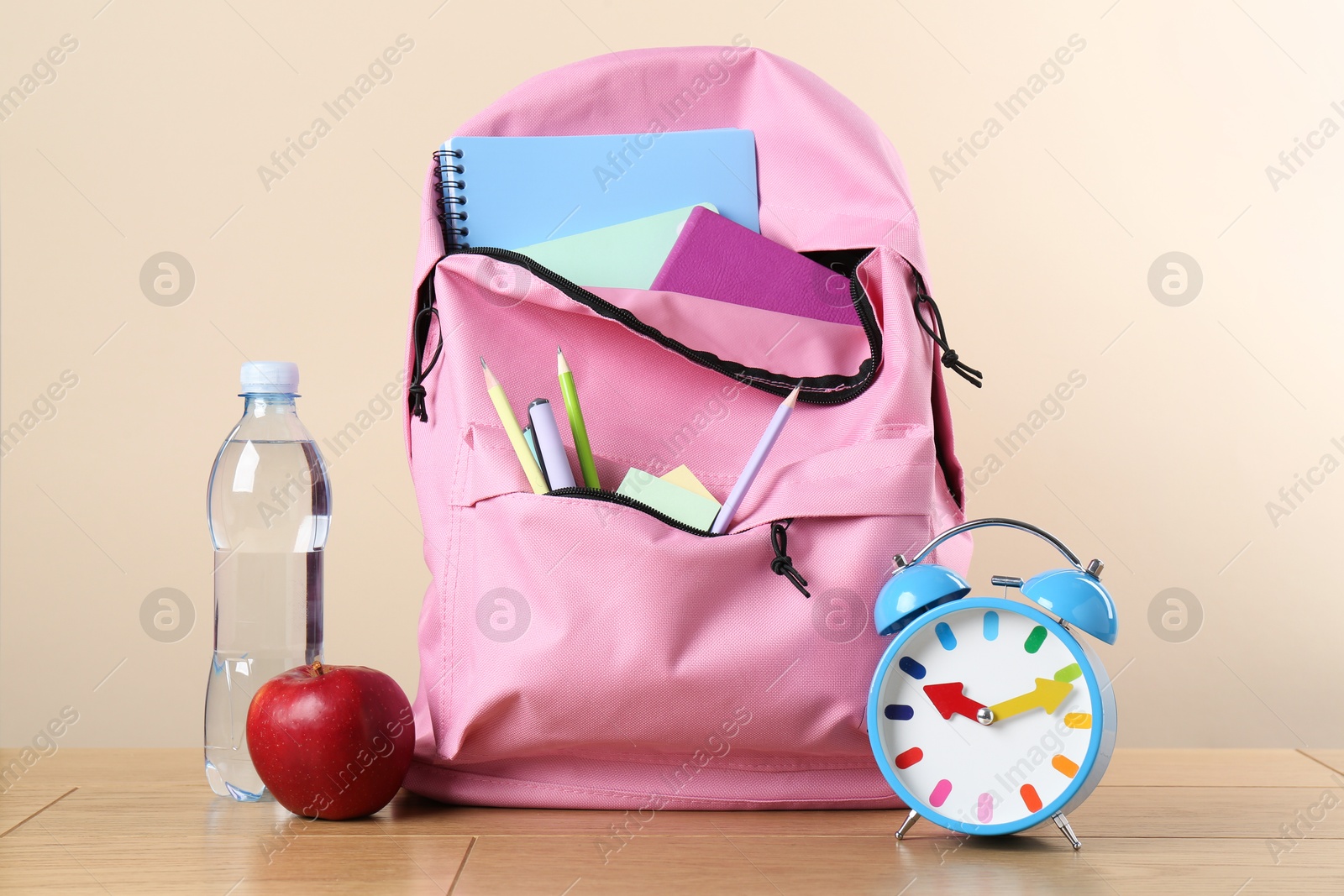 Photo of Backpack with different school stationery, alarm clock, bottle of water and apple on wooden table against beige background