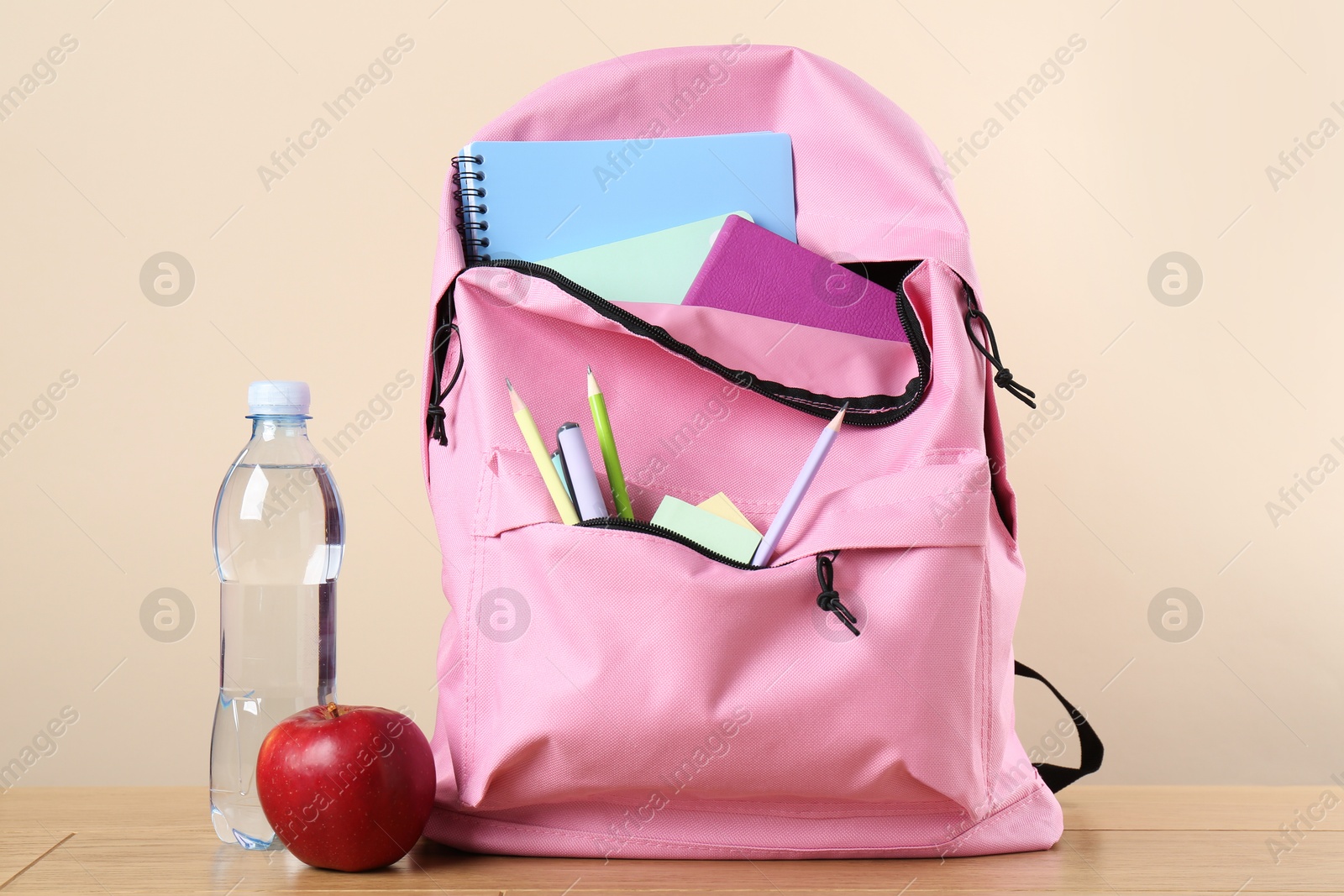 Photo of Backpack with different school stationery, bottle of water and apple on wooden table against beige background